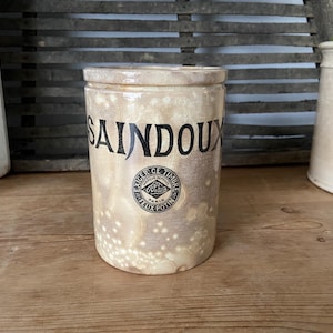 May include: A vintage ceramic jar with a white glaze and black lettering. The jar says "SAINDOUX" and has a black and white logo with the text "EXIGER CE TIMBRE" and "FELIX POTIN PARIS".