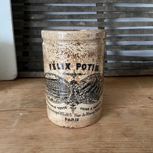 May include: A vintage ceramic jar with a brown and white speckled glaze. The jar has a black label with the text "Félix Potin" and details about the company's location and address in Paris, France.