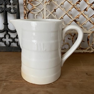 May include: A white ceramic jug with a handle. The jug has a textured surface and a slightly rounded shape.