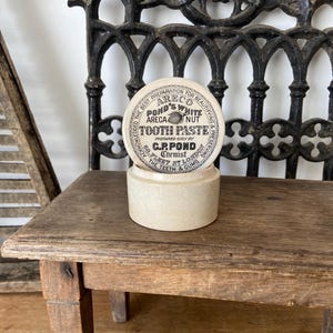 Könnte beinhalten: Ein zylindrischer, cremefarbener Keramiktopf mit einem Deckel, der den schwarzen Aufdruck trägt: ARECO POND'S WHITE ARECA NUT TOOTH PASTE PREPARED DAILY BY C.P. POND CHEMIST 68 FLEET ST LONDON THE TEETH & GUMS ACKNOWLEDGED THE BEST PREPARATION.