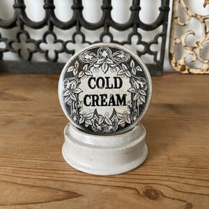 Könnte beinhalten: Ein rundes, weißes Keramikgefäß mit einem Deckel, der einen schwarz-weißen dekorativen Rand aus Blättern und Ranken aufweist. Der Text COLD CREAM ist in schwarzen, fetten Großbuchstaben in der Mitte des Deckels aufgedruckt. Der Sockel ist aus Keramik.
