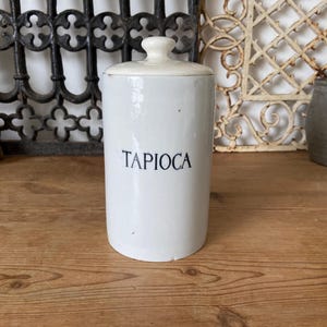 以下が含まれることがあります： 紺色の文字で「TAPIOCA」と書かれた白いセラミックジャー。ジャーは円筒形で、蓋には小さなノブが付いています。背景には装飾的な金属細工やその他のヴィンテージアイテムが含まれています。