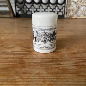 Puede incluir: Un pequeño tarro cilíndrico blanco con tapa, que presenta una etiqueta en blanco y negro con el texto La Reine des Cremes, Bossard-Lenoir, e incluye una ilustración de una persona y texto sobre su origen en Saint-Mande, París.