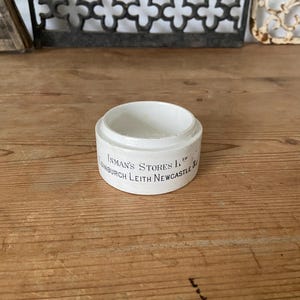 Op de afbeelding: Een kleine, cilindervormige witte keramische pot met een platte bodem en een open bovenkant. De buitenkant is voorzien van zwarte gedrukte tekst met de tekst Inman's Stores Ltd, Edinburgh, Leith, Newcastle. Het keramische oppervlak is glad en ondoorzichtig.