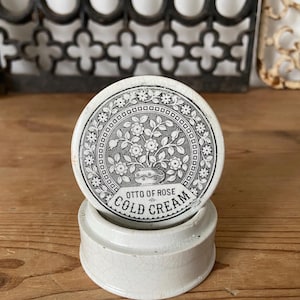 Könnte beinhalten: Ein runder weißer Keramiktopf mit einem Deckel, der eine schwarz-weiße Blumenillustration in einem kreisförmigen Rahmen zeigt. Der Text auf dem Deckel lautet OTTO OF ROSE COLD CREAM. Die Keramikoberfläche weist ein feines Craquelé-Muster auf.