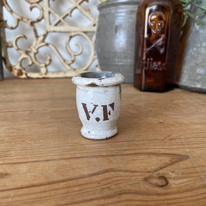 May include: A small, antique white ceramic jar with the letters "V.F." in brown. The jar has a distressed, aged appearance, with visible cracks in the glaze. The jar is on a wooden surface.