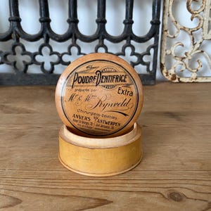 May include: A round, light-colored wooden container with a lid featuring black printed text that reads Poudre Dentifrice, Extra, Mr & Mme Rynveld, Chirurgiens-Dentistes, Anvers, Antwerpen, and additional address details in a vintage style.
