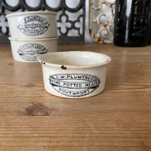 Puede incluir: Tres tarros de cerámica de color blanco roto con un acabado de esmalte agrietado. Cada tarro presenta una impresión ovalada negra que dice G.W. PLUMTREE HOME POTTED MEATS SOUTHPORT. Los tarros tienen una forma cilíndrica baja con un pequeño borde y desgaste visible.