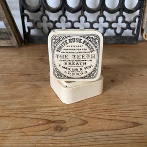 May include: A vintage off-white square container with a lid featuring black text that reads White Rose Paste, An Elegant Preparation For Preserving & Whitening The Teeth, Purifying & Perfuming The Breath, Prepared By S. Maw, Son & Sons, London.