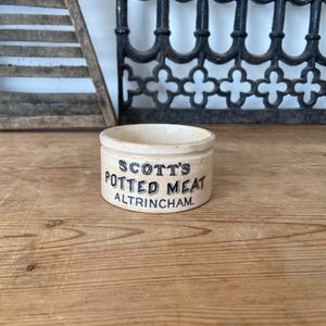 Puede incluir: Un pequeño recipiente cilíndrico de cerámica de color crema con un borde plano. El exterior presenta un texto impreso en negro que dice SCOTT'S POTTED MEAT ALTRINCHAM. El recipiente tiene un acabado liso y esmaltado con un diseño sencillo y funcional.