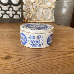 May include: A vintage, round Borax container with a lid. The container is white with blue lettering and a design that reads "Home Sweet Home." The lid has blue text that reads "Borax Co." The container is on a wooden surface.