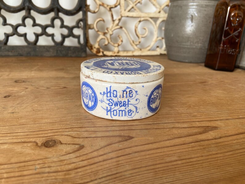 May include: A vintage, round Borax container with a lid. The container is white with blue lettering and a design that reads "Home Sweet Home." The lid has blue text that reads "Borax Co." The container is on a wooden surface.