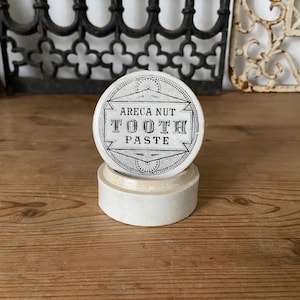 May include: A round, off-white container with a lid featuring black printed text that reads ARECA NUT TOOTH PASTE. The container has a smooth, matte finish and a cylindrical base, displaying a vintage-style design with geometric border details.
