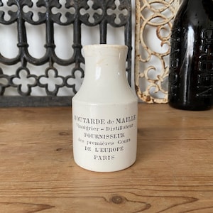 Puede incluir: Un tarro de mostaza de cerámica blanca con cuello estrecho y base cilíndrica. El frente presenta un texto impreso en negro que dice MOUTARDE de MAILLE Vinaigrier - Distillateur FOURNISSEUR des premières Cours DE L'EUROPE PARIS.