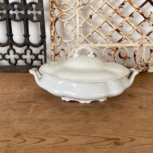 May include: White porcelain serving dish with a lid and scalloped edges. The dish is on a wooden surface.