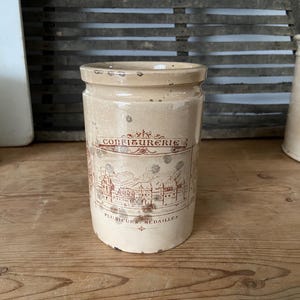 May include: A vintage beige ceramic jar with a cylindrical shape. The jar features a decorative label with the word "CONFITUREERIE" and a detailed illustration of a town. The jar is likely used for storing food items.