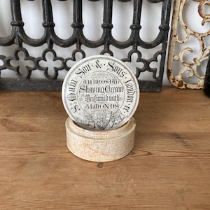 May include: A round ceramic shaving cream container with a cream-colored base featuring a fine crackle glaze. The lid displays black printed text reading S. Oam Son & Sons London, Ambrosial Shaving Cream, Perfumed with Almonds, and a crest.