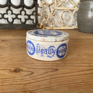 May include: A small, round, antique ceramic jar with a lid. The jar is off-white with blue lettering that reads "Borax Beauty" and "London". The lid also has blue text. The jar sits on a wooden surface.