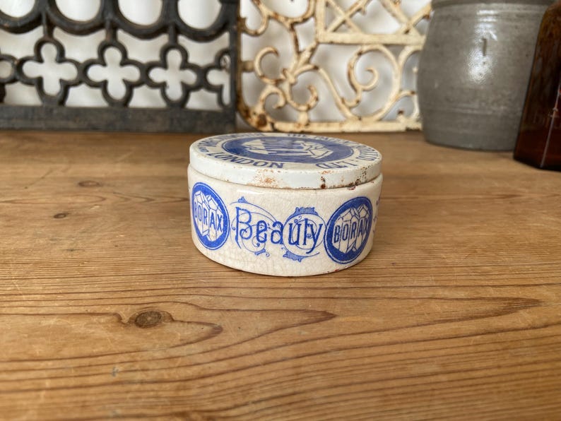 May include: A small, round, antique ceramic jar with a lid. The jar is off-white with blue lettering that reads "Borax Beauty" and "London". The lid also has blue text. The jar sits on a wooden surface.