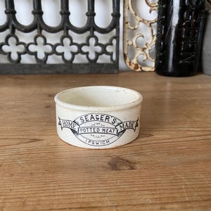 Puede incluir: Un recipiente de cerámica redondo de color blanco roto con una etiqueta impresa en negro que dice Seager's Home Made Potted Meat Ipswich. El envase tiene un acabado liso ligeramente desgastado y una forma cilíndrica baja.