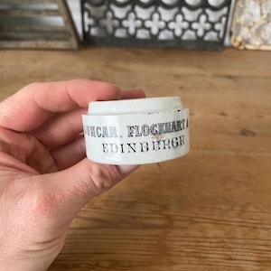 May include: A small, cylindrical white ceramic pot with a stepped rim. The side features black printed text that reads DUNCAN, FLOCKHART & CO. EDINBURGH. The surface shows minor signs of wear and discolouration along the edges of the lettering.