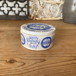 May include: A vintage, round Borax tin with a white and blue design. The lid features text that reads "BIRMINGHAM AND LONDON". The side of the tin has the words "Utility, Purity, Safety" in a decorative font. The tin is on a wooden surface.