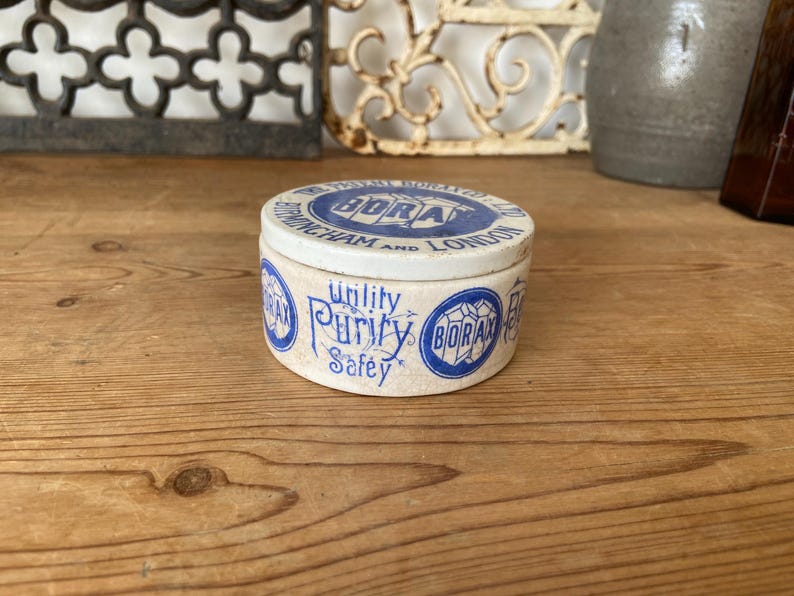 May include: A vintage, round Borax tin with a white and blue design. The lid features text that reads "BIRMINGHAM AND LONDON". The side of the tin has the words "Utility, Purity, Safety" in a decorative font. The tin is on a wooden surface.