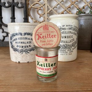 Puede incluir: Una colección de frascos y recipientes antiguos de la marca Keiller. El frasco central muestra "Keiller Raspberry Jam" con una etiqueta roja y verde. Otros frascos incluyen "Keiller & Sons Dundee Marmalade".