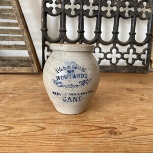 May include: A beige stoneware mustard pot with a rounded body and a short neck. The front features blue printed text that reads FABRIQUE DE MOUTARDE, P. Cierenteyn-Verlen, MARCHE AUX LEGUMES, GAND. The pot has a speckled texture and a smooth finish.