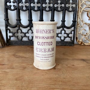 Könnte beinhalten: Ein zylindrischer, cremefarbener Behälter mit abgenutzter Oberfläche und dunkelroter Aufschrift HORNER'S DEVONSHIRE CLOTTED CREAM sowie kleinerem gedrucktem Text mit Angaben zur Beschaffung und dem Gründungsjahr 1898.