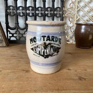 May include: A vintage ceramic mustard jar, cylindrical in shape. It's cream-coloured with blue stripes and the text "Moutarde Imperial." The jar sits on a wooden surface.