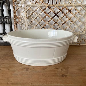 May include: White ceramic oval planter with two handles. The planter has a ribbed design and is sitting on a wooden surface.