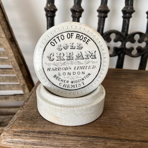 Könnte beinhalten: Ein runder, cremefarbener Keramiktopf mit einem Deckel, der den schwarzen Aufdruck OTTO OF ROSE COLD CREAM HARRODS LIMITED LONDON BECHER WIGGINTON CHEMIST trägt. Die Keramikoberfläche weist durchgehend eine feine, gealterte Craquelé-Glasur auf.