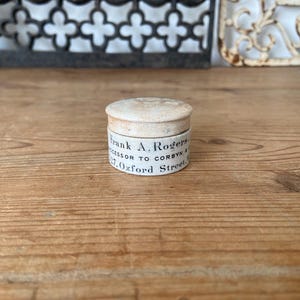 Op de afbeelding: Een kleine, ronde, gebroken witte container met deksel en een verweerde textuur. De zijkant bevat zwarte gedrukte tekst met de tekst Frank A. Rogers, Successor to Corbyn, 27 Oxford Street. Het deksel heeft een licht gewelfde, matte afwerking.