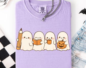May include: A light purple t-shirt featuring a graphic of four cartoon ghosts, each holding a different item: a pencil, a book, a mug, and a jack-o'-lantern. The shirt has a Comfort Colors tag and a silver chain necklace.