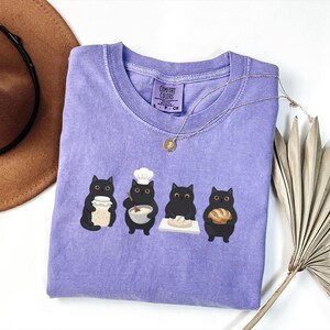 May include: A light purple short-sleeve t-shirt with a graphic print of four black cats baking. The cats are shown holding a jar, mixing in a bowl while wearing a chef hat, kneading dough on a board, and holding a loaf of bread. The shirt has a Comfort Colors label.