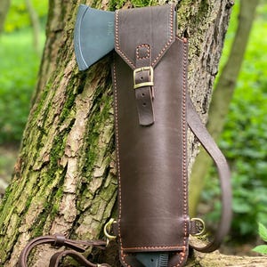 May include: A dark brown leather axe sheath with a matching shoulder strap and a small axe. The sheath has a buckle closure and is stitched with orange thread. The axe head is gray.