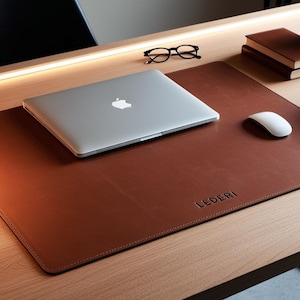 Puede incluir: Un tapete de escritorio de cuero marrón con la palabra "LEDERI" grabada en relieve. Una computadora portátil plateada, un ratón blanco y gafas están sobre el tapete. Libros y una pequeña planta en maceta también están en el escritorio de madera.