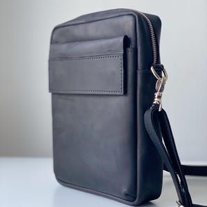 May include: Black leather crossbody bag with a rectangular shape. Features a front flap pocket, a top zipper closure, and a detachable shoulder strap. The bag has silver-tone hardware and a minimalist design.