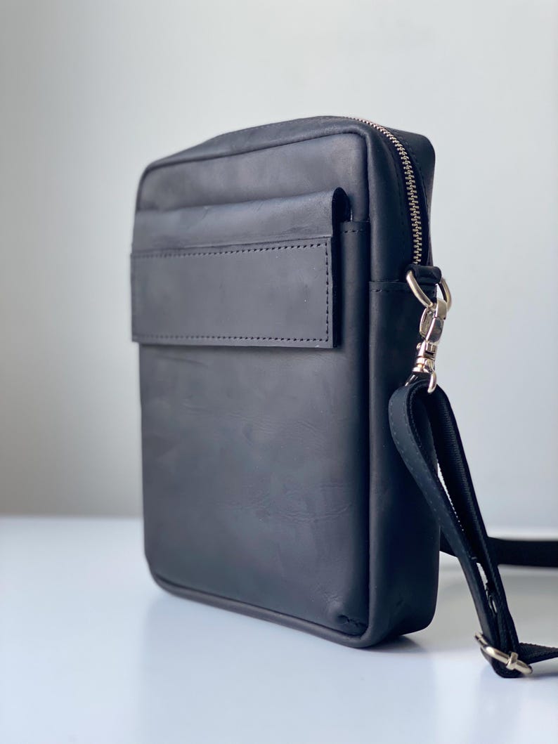 May include: Black leather crossbody bag with a rectangular shape. Features a front flap pocket, a top zipper closure, and a detachable shoulder strap. The bag has silver-tone hardware and a minimalist design.
