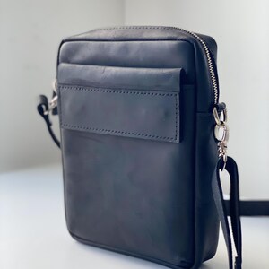 May include: Black leather crossbody bag with a rectangular shape. The bag features a front pocket, a top zipper closure, and a silver-tone metal strap attachment. The adjustable strap is black.