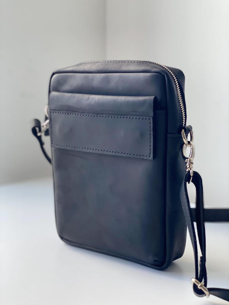 May include: Black leather crossbody bag with a rectangular shape. The bag features a front pocket, a top zipper closure, and a silver-tone metal strap attachment. The adjustable strap is black.