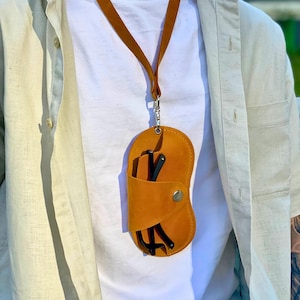 Può includere: Un astuccio per occhiali in pelle giallo senape con un cordino abbinato. La custodia è di forma ovale con chiusura a scatto e contiene occhiali neri. Il cordino è attaccato con un fermaglio argentato. La custodia si indossa al collo.
