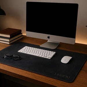 May include: A black leather desk mat with the word "LEDERJ" embossed in white. The mat is on a wooden desk with a computer, keyboard, mouse, glasses, and a stack of books.