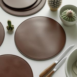 May include: A collection of brown leather placemats arranged on a white surface. The round placemats are of varying sizes. Two knives with wooden handles and a white bowl are also present. Small cacti in pots add a touch of greenery to the scene.