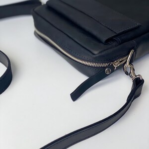 May include: A black leather crossbody bag with a rectangular shape. The bag features a silver zipper, a small front pocket, and a long, adjustable strap. The bag is designed for carrying essentials.