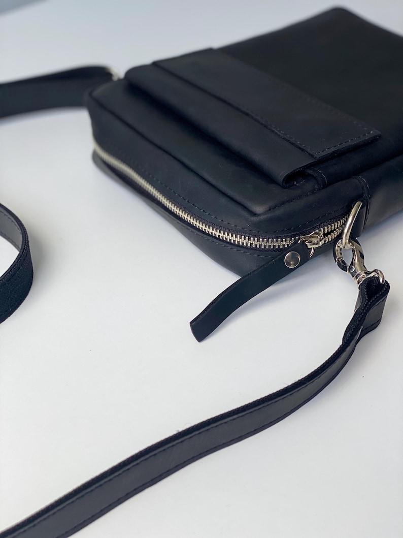 May include: A black leather crossbody bag with a rectangular shape. The bag features a silver zipper, a small front pocket, and a long, adjustable strap. The bag is designed for carrying essentials.