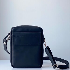 May include: Black leather crossbody bag with a rectangular shape. The bag features a front pocket, a long adjustable strap, and silver-tone hardware. The bag is designed for everyday use and has a minimalist aesthetic.