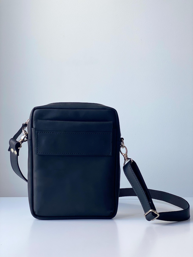 May include: Black leather crossbody bag with a rectangular shape. The bag features a front pocket, a long adjustable strap, and silver-tone hardware. The bag is designed for everyday use and has a minimalist aesthetic.