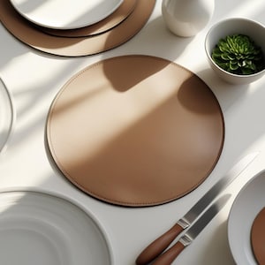 May include: A tan leather placemat with a stitched border is the focal point of this image. White plates, a small white vase, a bowl with a green succulent, and two knives with wooden handles are also visible. The items are arranged on a white surface.